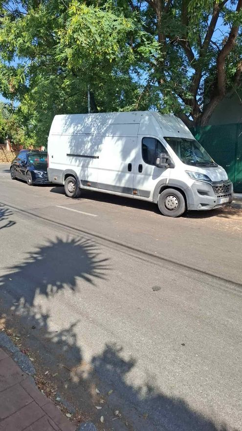 Una furgoneta blanca estacionada en la calle junto a un árbol. Se ve un coche negro detrás.