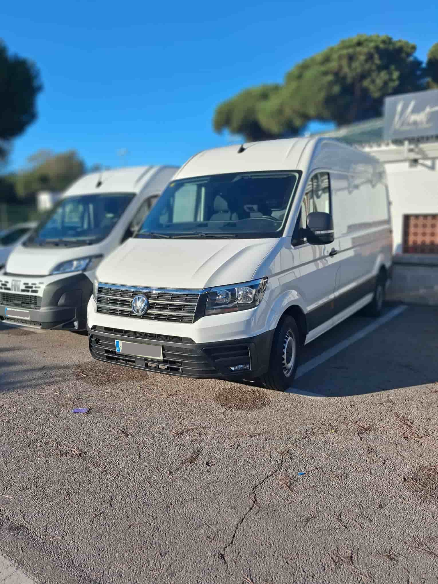 Furgoneta Volkswagen Crafter blanca estacionada sobre grava frente a un edificio; otra furgoneta a la izquierda.