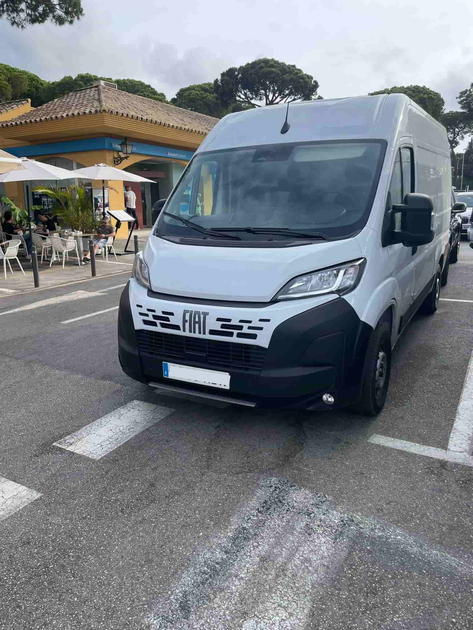 Furgoneta Fiat Ducato blanca estacionada en una carretera, cerca de un restaurante con mesas al aire libre.