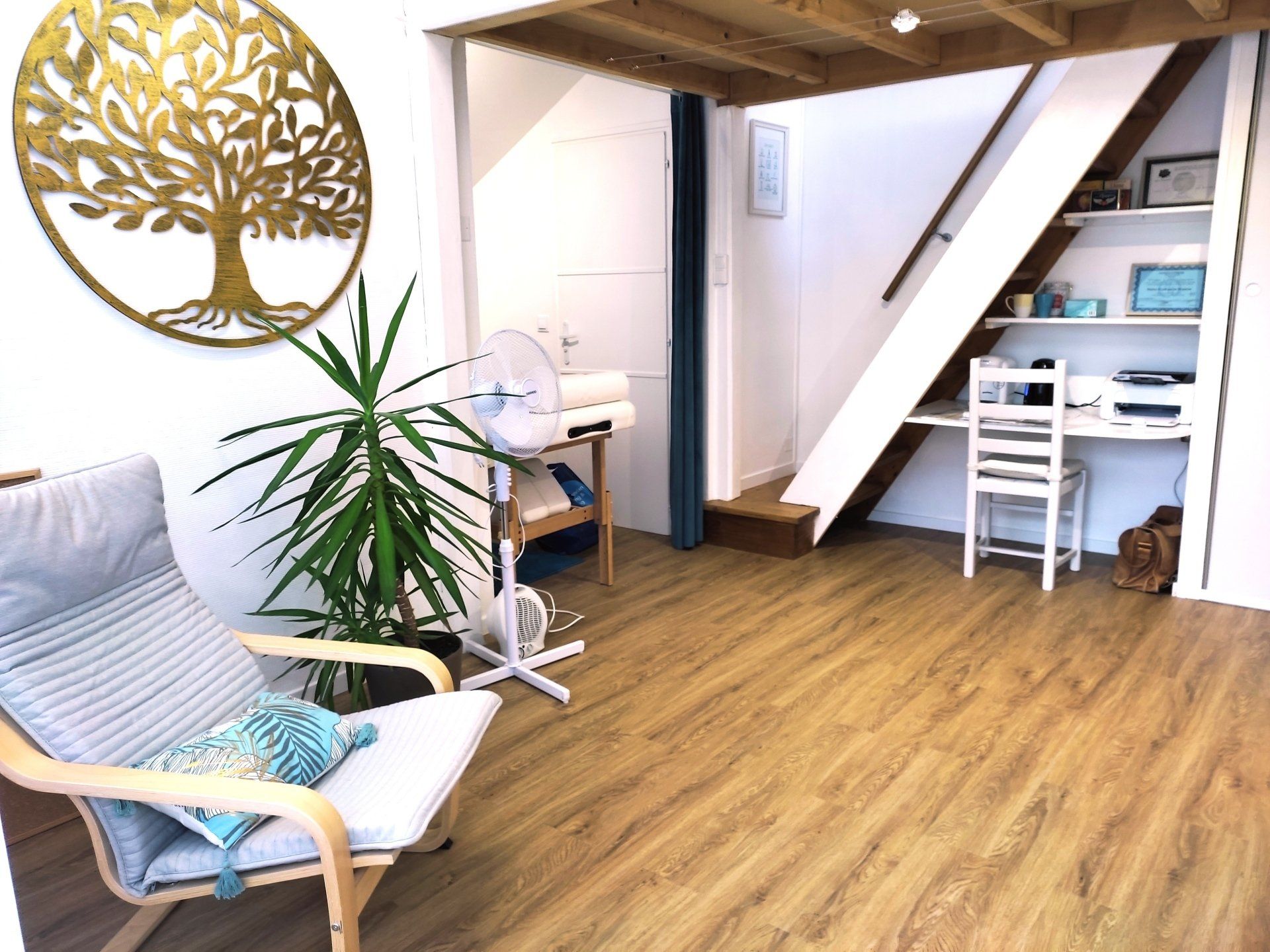 Intérieur chaleureux avec chaise, bureau, grand arbre et ventilateur. Parquet et escalier en bois. Luminosité naturelle.