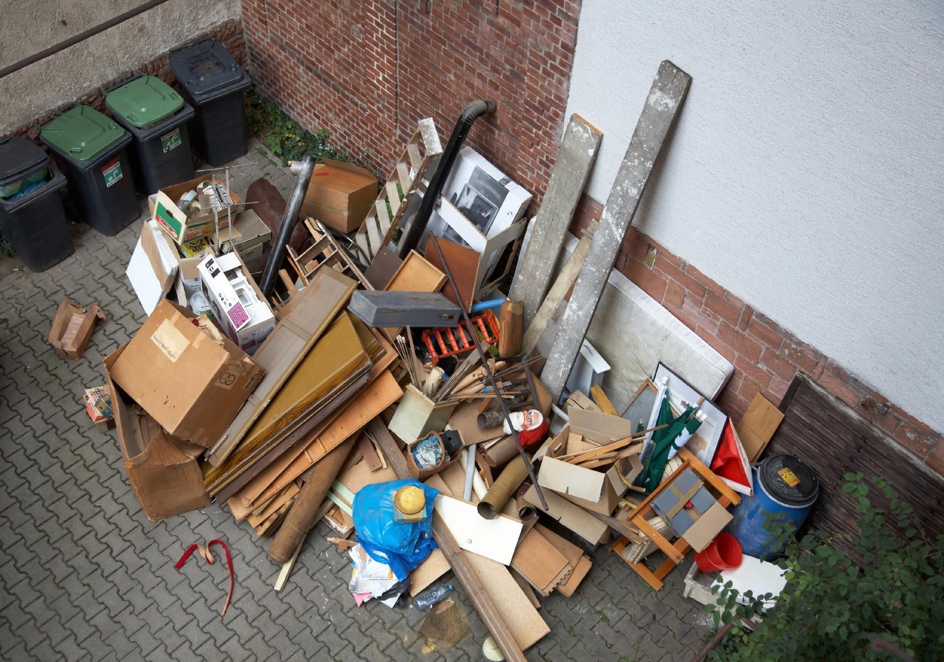 Überfüllter Stapel aus Pappe und Holzabfällen neben einer Backsteinmauer und Mülltonnen, von oben gesehen.