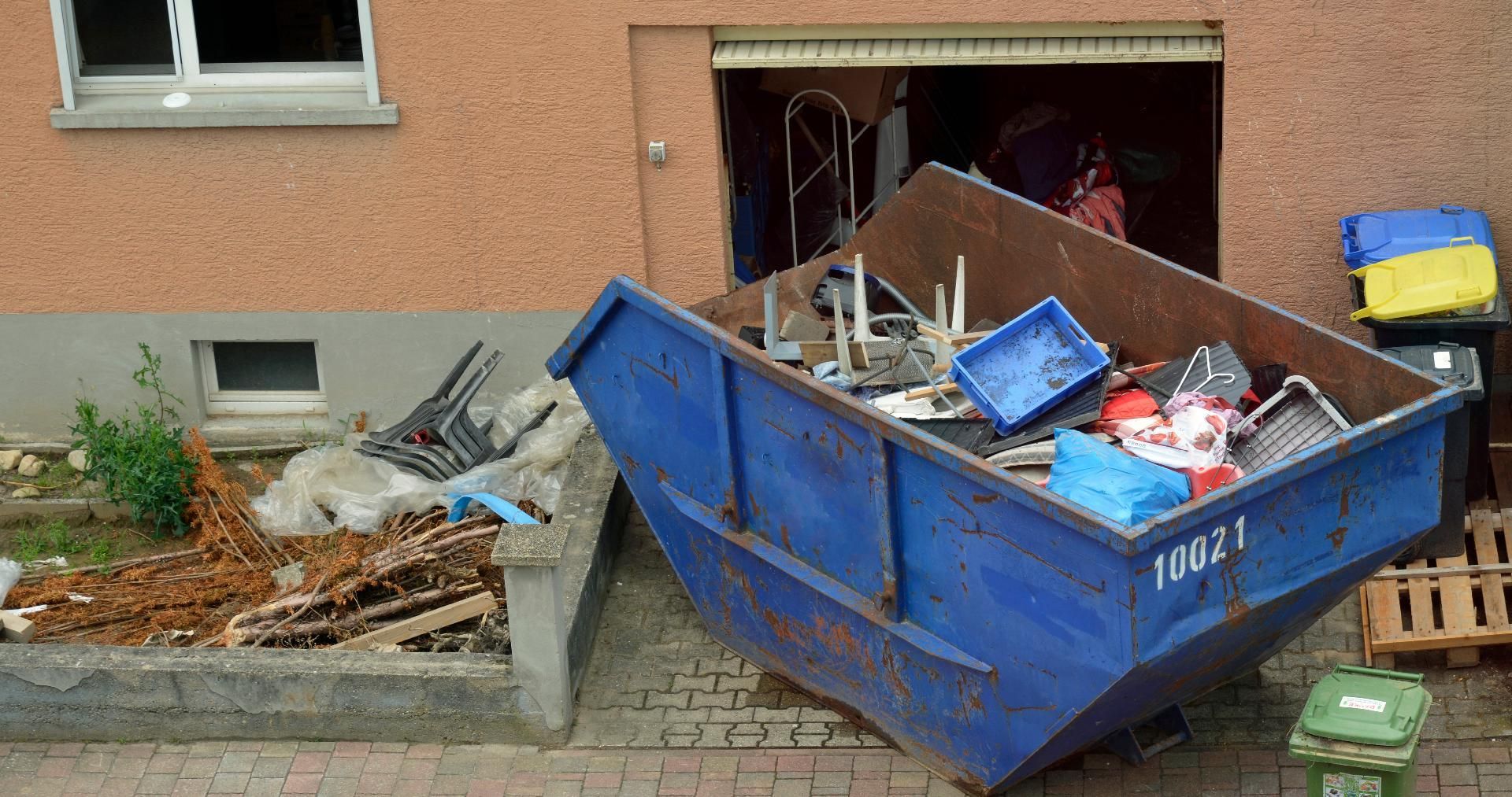 Ein blauer Müllcontainer, der neben einem Gebäudeeingang und einer Mauer überquillt, ist voller Bauschutt.