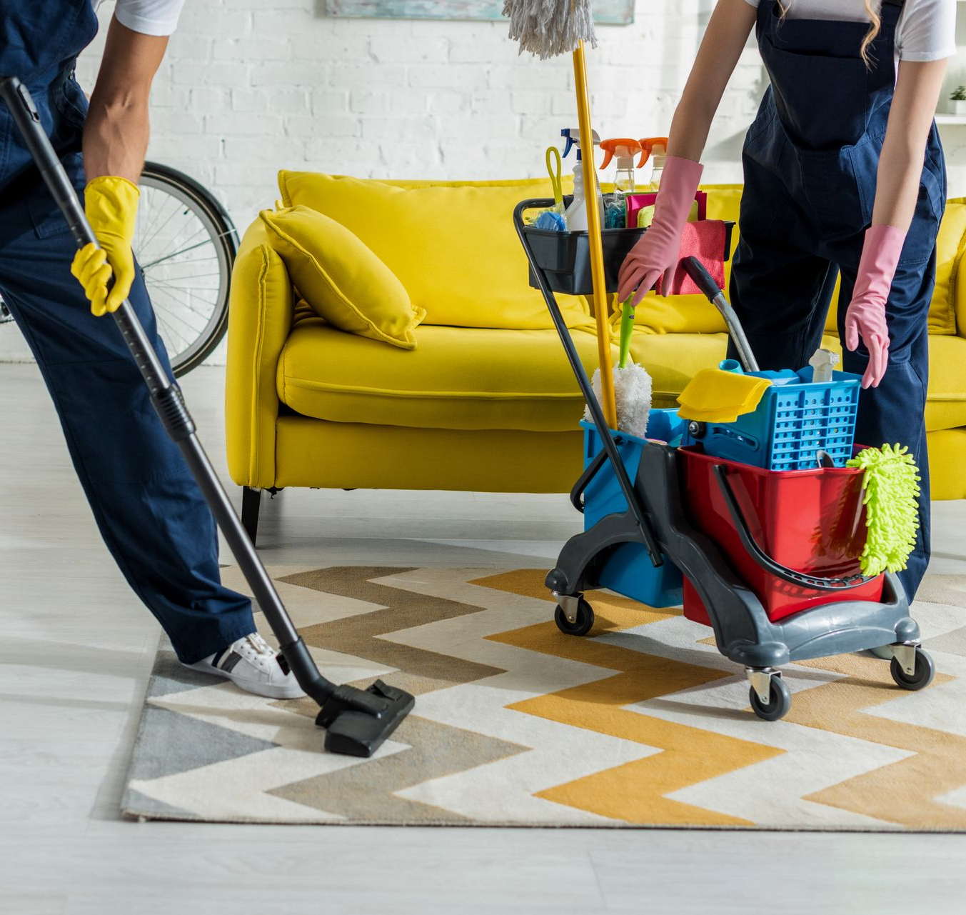 Deux personnes nettoient le salon avec un aspirateur et un chariot de nettoyage ; canapé jaune, tapis.