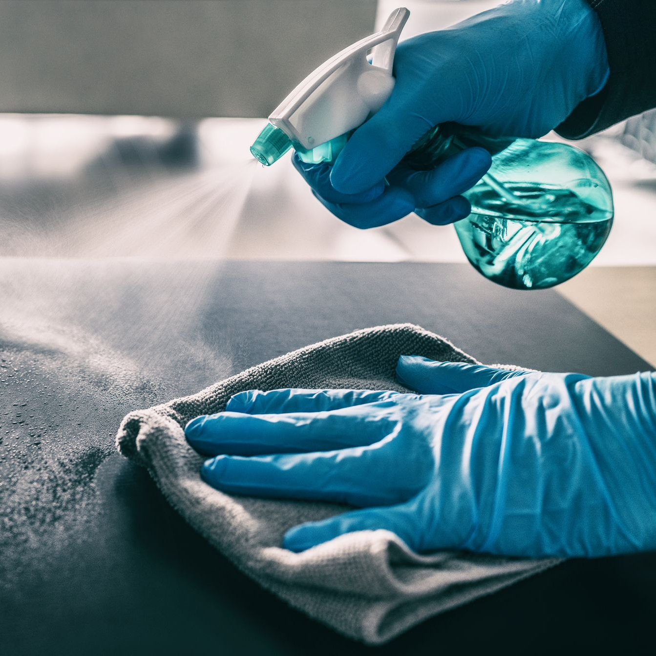 Une personne portant des gants bleus vaporise et essuie une surface avec des produits de nettoyage.