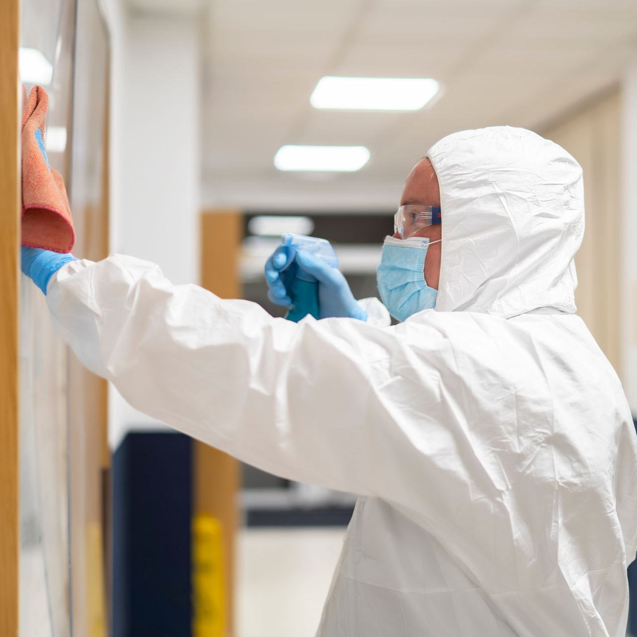 Une personne en combinaison de protection blanche vaporise et essuie un mur dans un couloir.