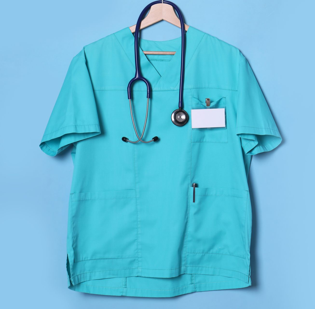 Blouse médicale turquoise avec stéthoscope et étiquette nominative vierge, accrochée à un mur bleu.