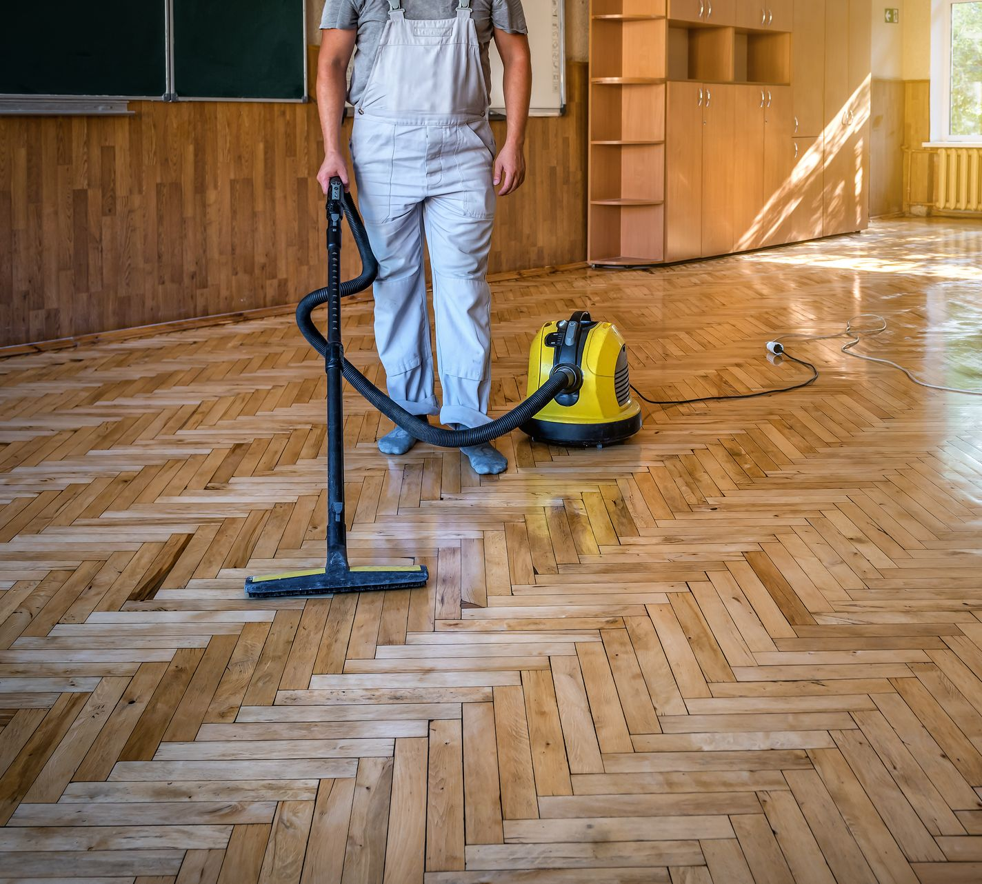 Une personne en salopette passe l'aspirateur sur un parquet à chevrons dans une pièce avec un tableau noir et des armoires en bois.