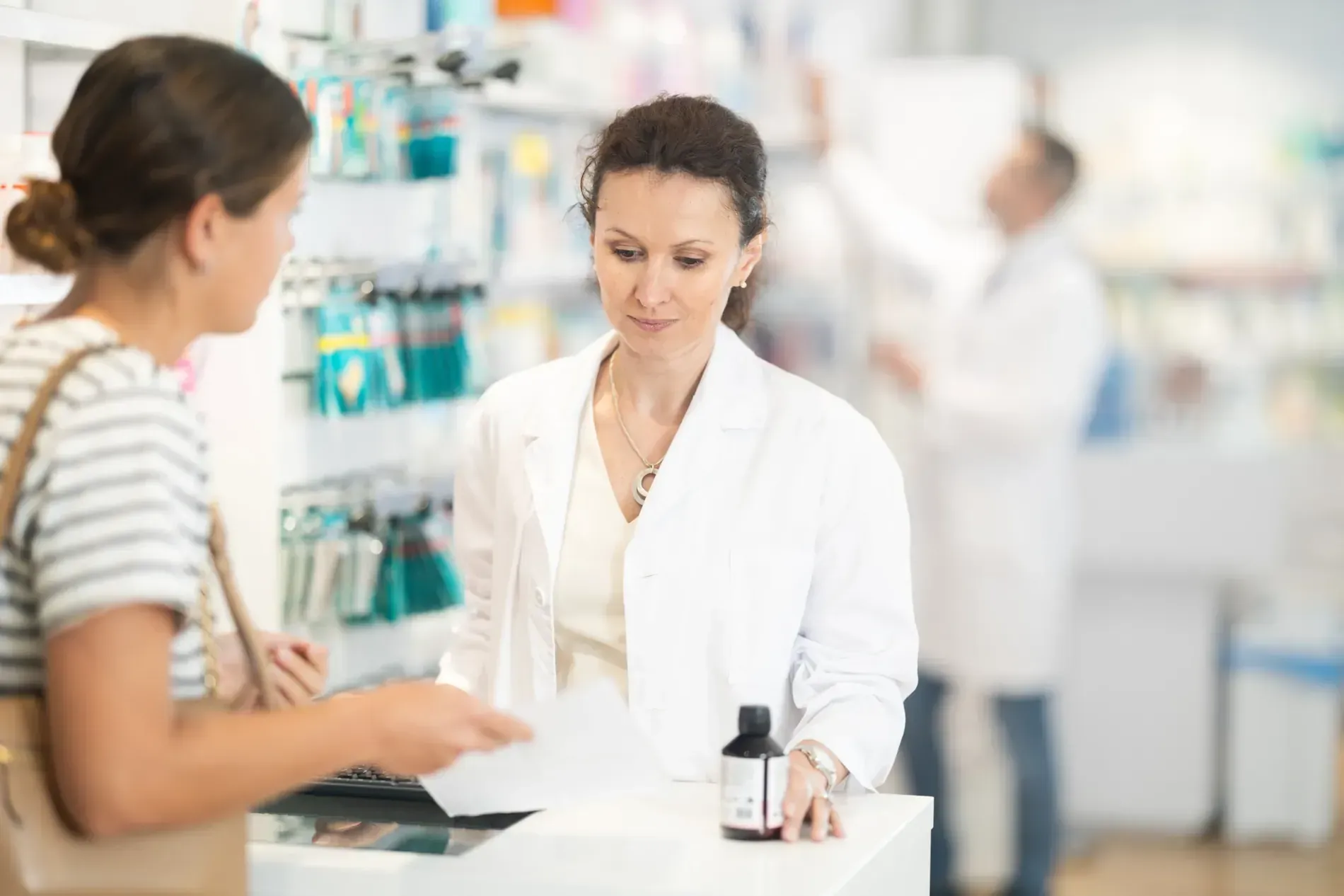 Mujer con bata de farmacia atendiendo a un cliente, con una botella de medicamento en el mostrador.