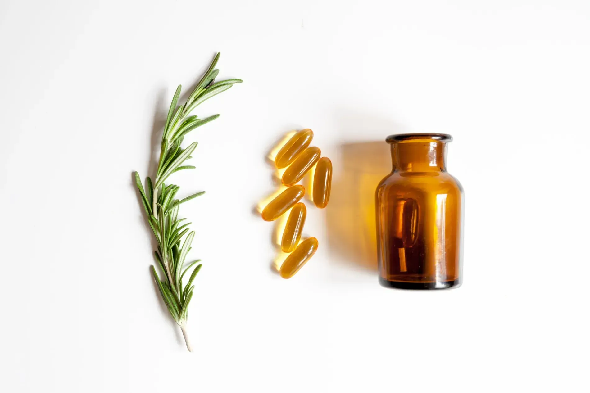 Ramita de romero, cápsulas amarillas y pequeña botella de vidrio marrón sobre superficie blanca.