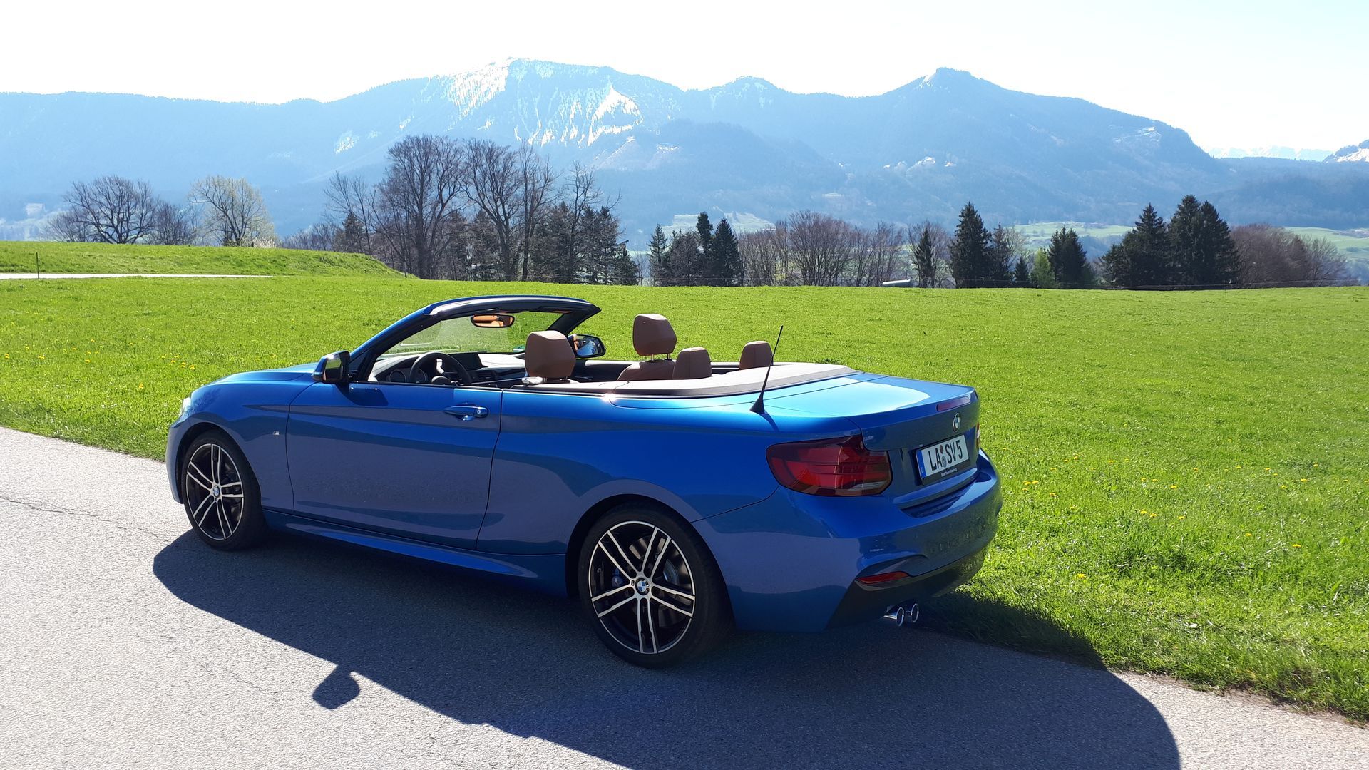 Blauer Cabrio-Pkw auf Landstraße vor grüner Wiese und Bergen im Hintergrund