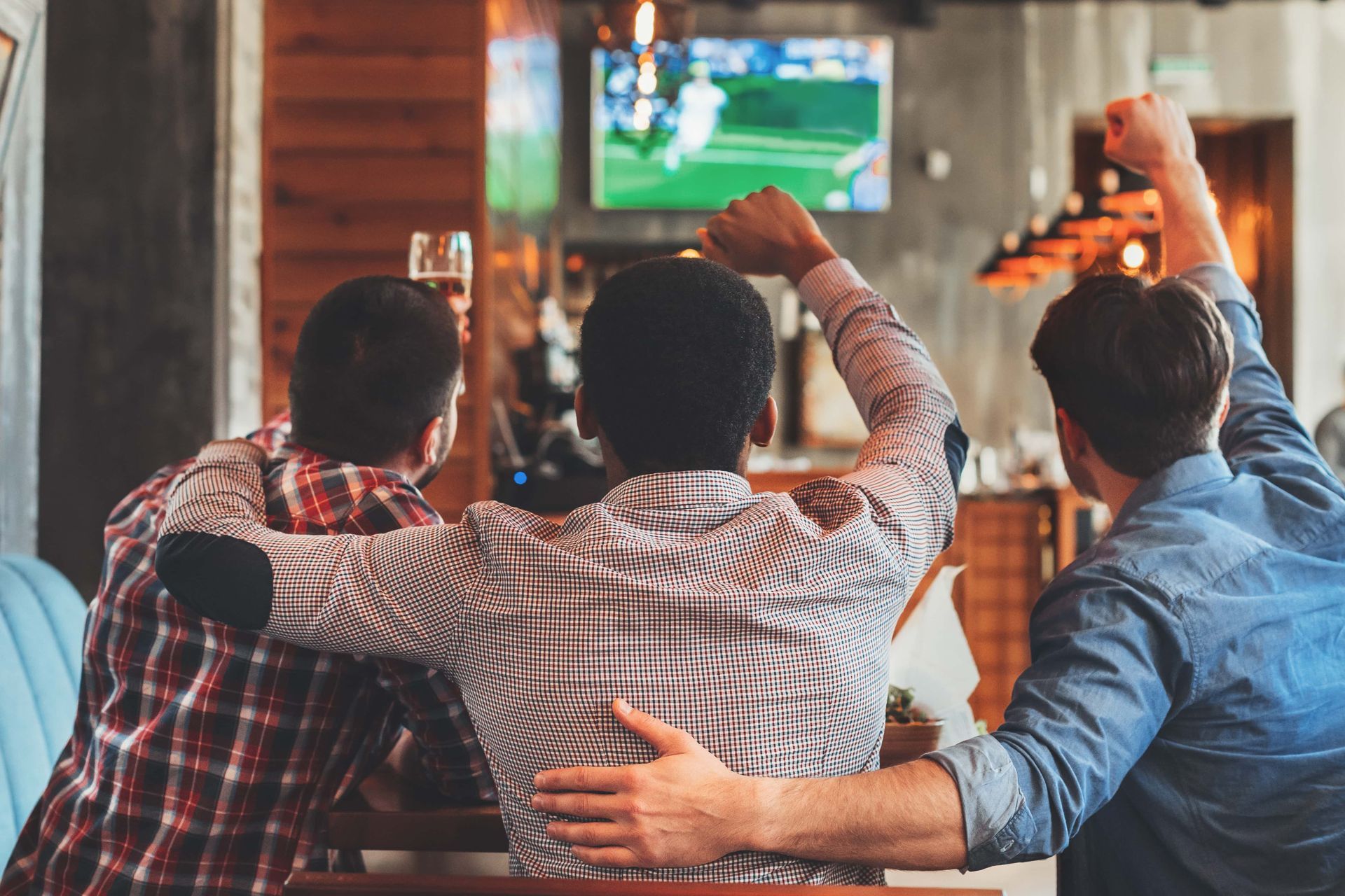 Trois amis, assis sur un canapé, applaudissent devant la télévision dans un bar chaleureux, leurs verres levés.
