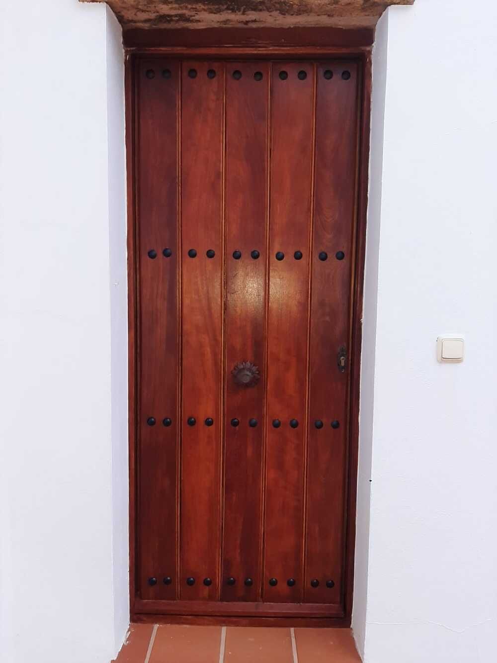 Puerta de madera con paneles de color marrón oscuro, rematada con remaches negros, colocada en una pared blanca.