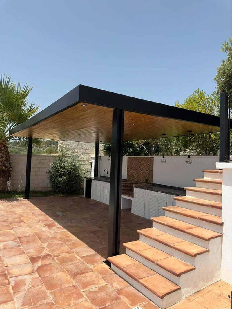 Patio con toldo negro, escalones, piso de ladrillo y cocina al aire libre en un día soleado.