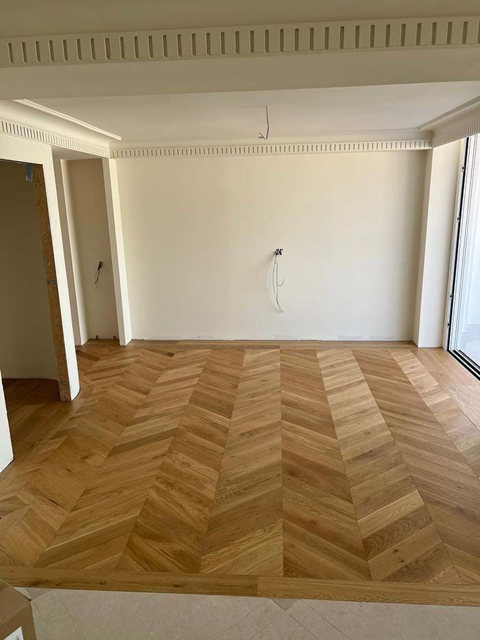 Habitación vacía con suelo de parqué de madera clara con diseño de chevrones. Paredes blancas, molduras decorativas en el techo y una puerta.