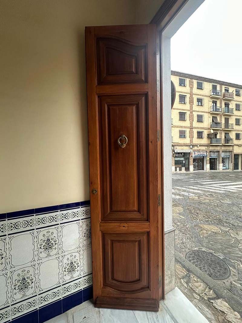 Puerta de madera con paneles decorativos y llamador de latón, abierta a un patio de piedra y edificios de la ciudad.