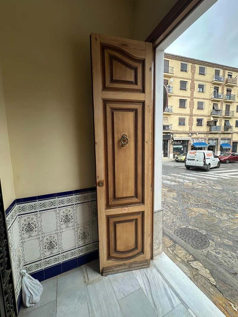 Puerta de madera en una entrada abierta, paneles ornamentados, llamador de latón, escena callejera visible.