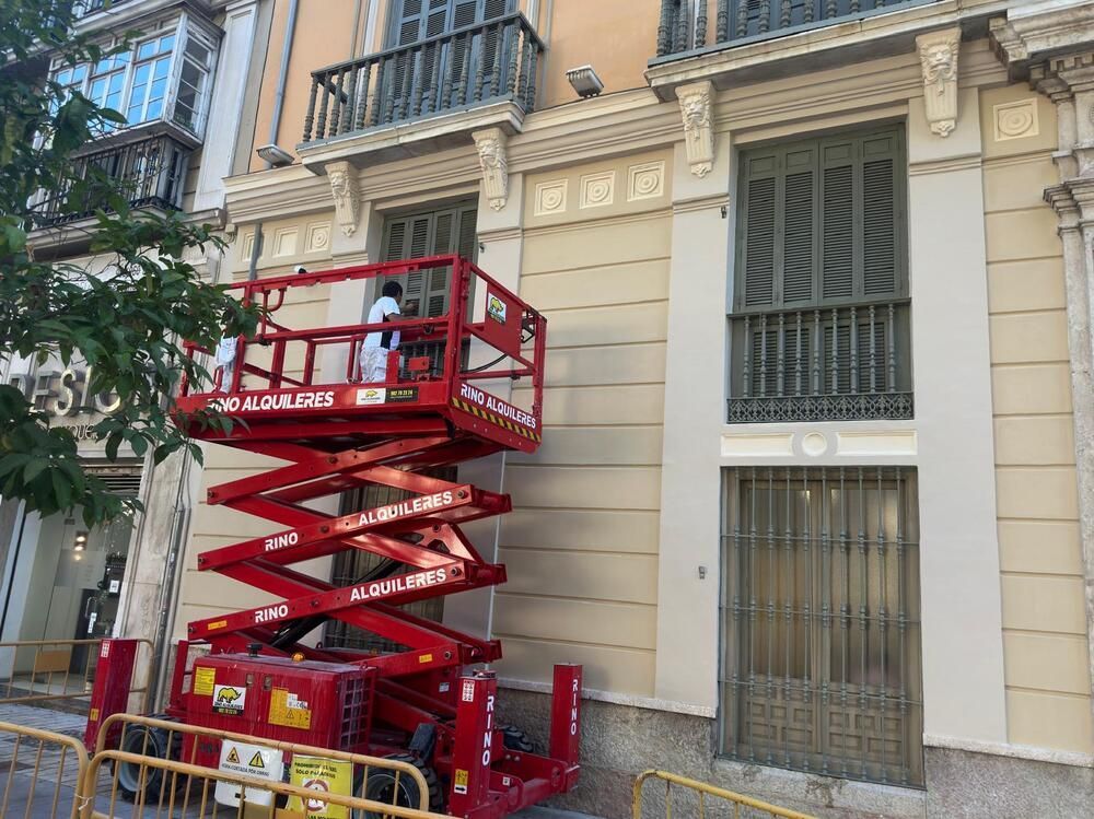 Trabajadores en un elevador de tijera rojo pintando la fachada de un edificio con persianas.