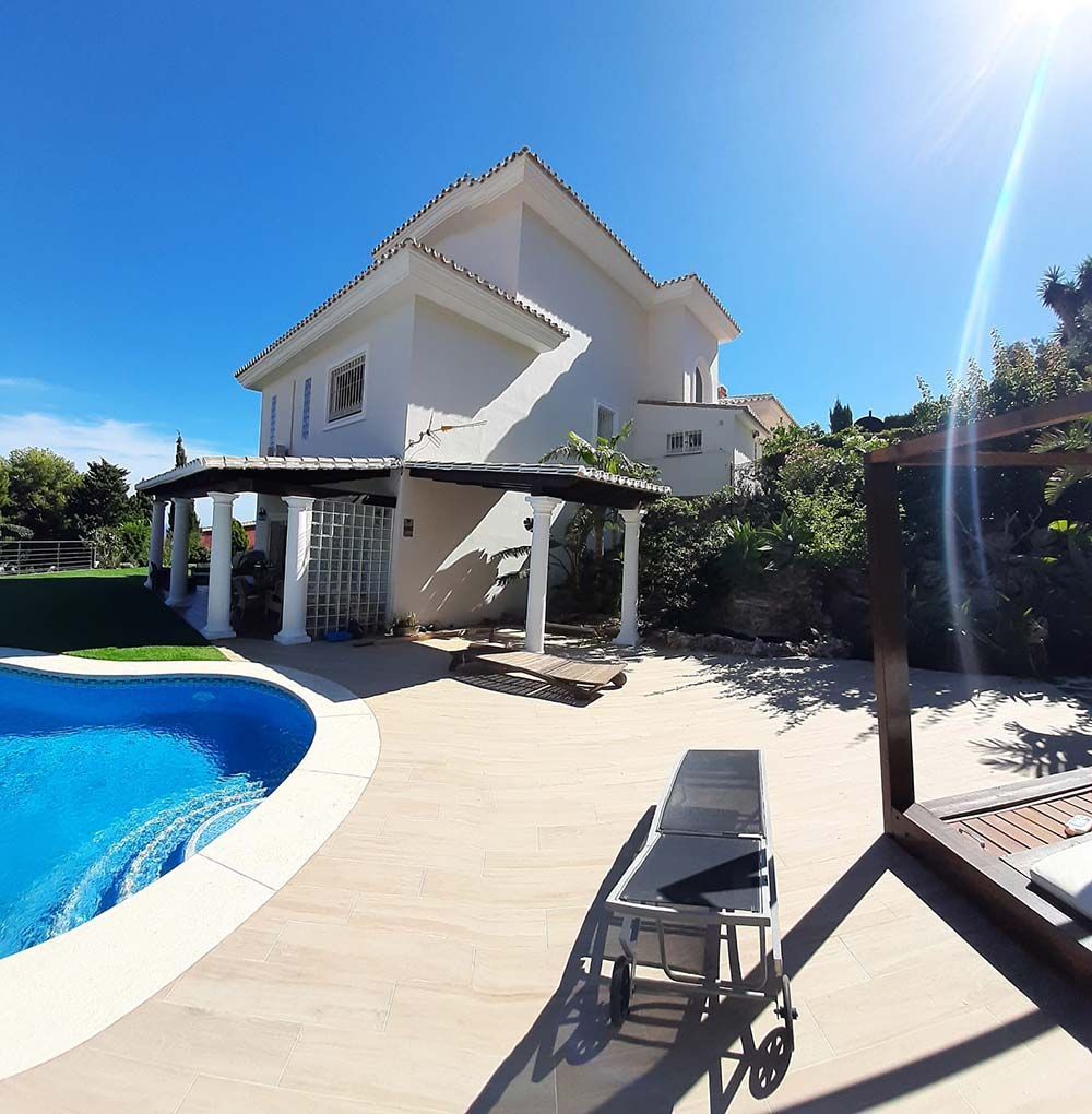 Casa blanca de dos plantas con piscina y pérgola. Cielo azul brillante. Terraza de madera con tumbona.