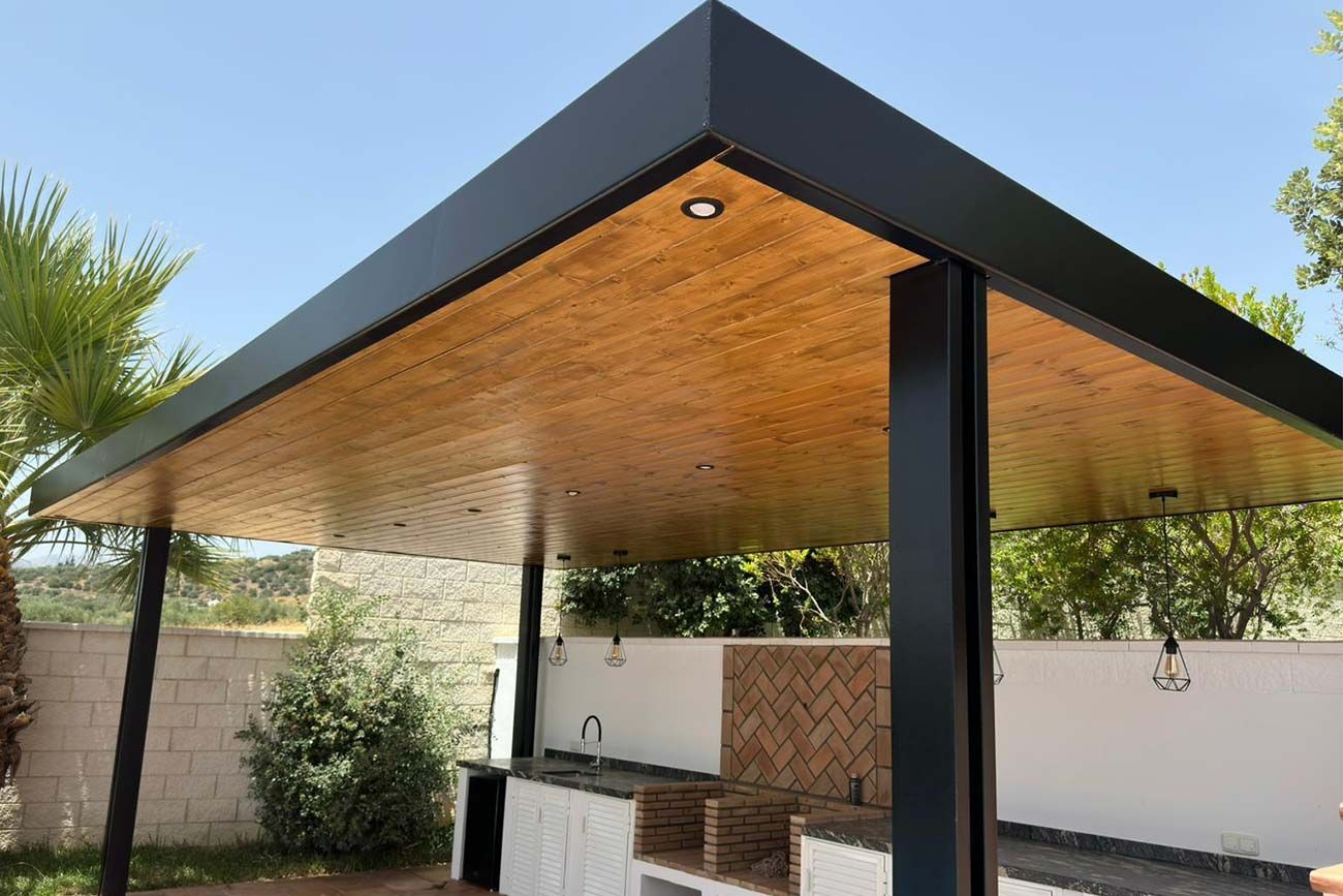 Pérgola exterior con marco negro, techo de madera, cocina incorporada y luces de cadena, frente a un cielo soleado y un patio.