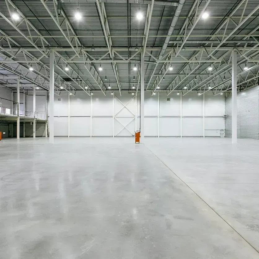 Gran almacén industrial vacío con suelo de hormigón, paredes blancas y techo de metal con luces brillantes.