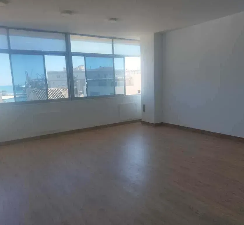 Habitación vacía con grandes ventanales, suelo de madera y paredes blancas. Vista exterior soleada durante el día.