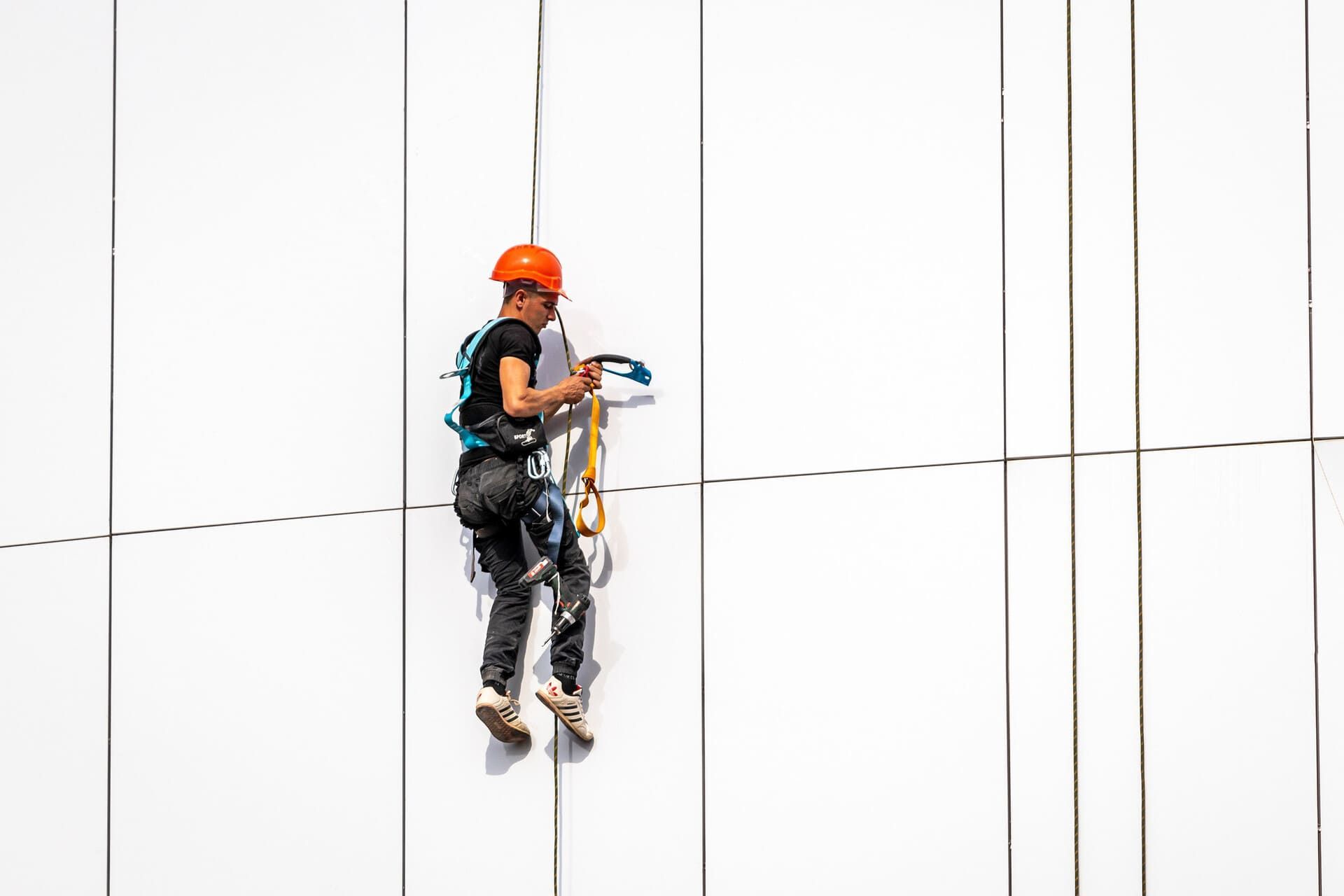 Persona con casco naranja suspendida en la pared blanca de un edificio, limpiando o inspeccionando, mirando hacia atrás.