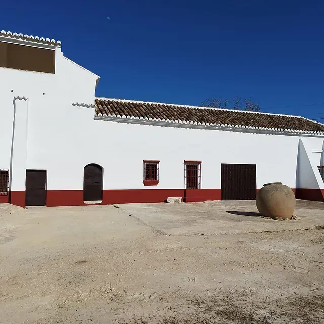 Edificio blanco con detalles rojos, puertas marrones y una gran olla de barro en un patio bajo un cielo azul.