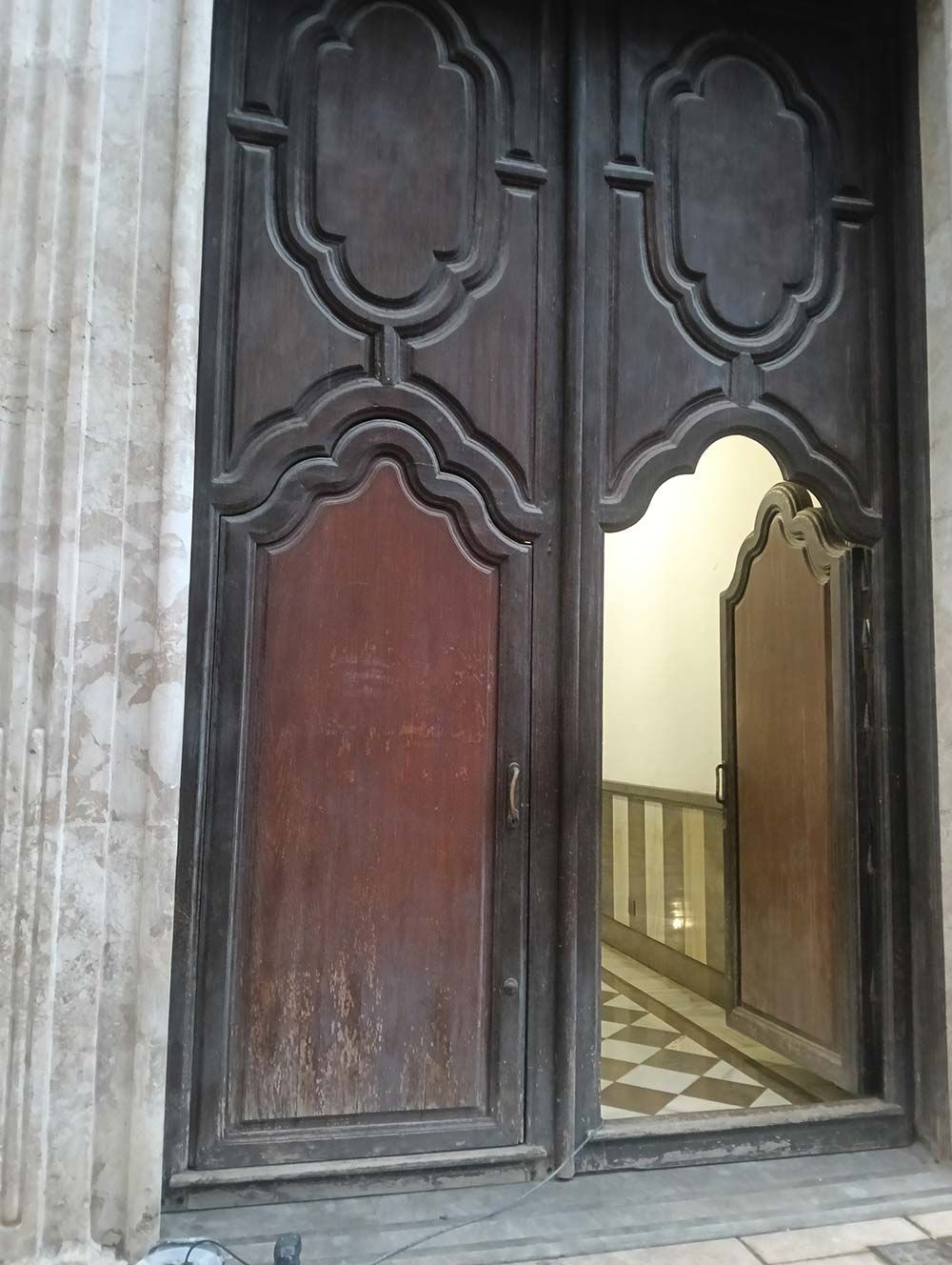 Las puertas dobles de madera, parcialmente abiertas, revelan un pasillo con piso cuadriculado.