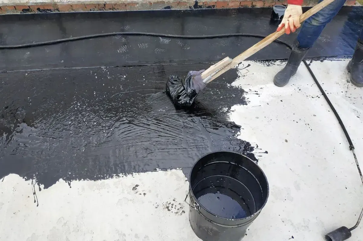 Persona esparciendo alquitrán negro sobre un techo plano con un cepillo y un balde.