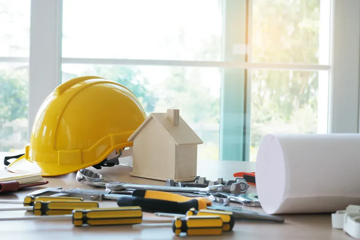Casco amarillo, maqueta de casa y herramientas sobre una mesa delante de una ventana.