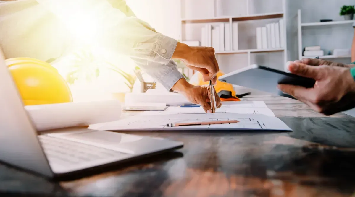 Dos personas revisando planos en un escritorio con una computadora portátil, una tableta y un casco de construcción.