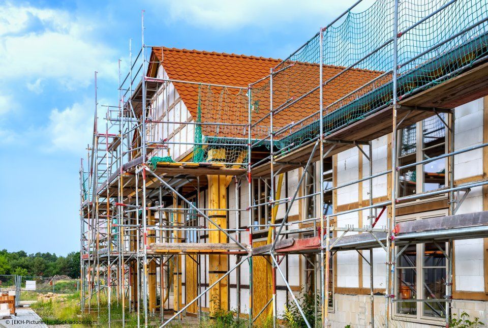Ein im Bau befindliches Fachwerkgebäude, dessen Außenfassade von einem Gerüst verdeckt wird, vor blauem Himmel.