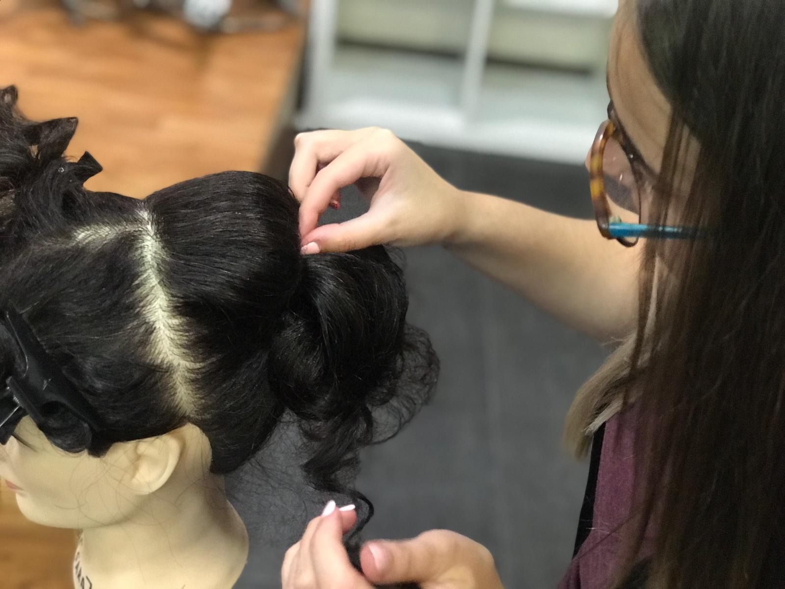 Una persona peina a un maniquí de cabello oscuro, sujetándole el cabello rizado y usando anteojos en un entorno iluminado.