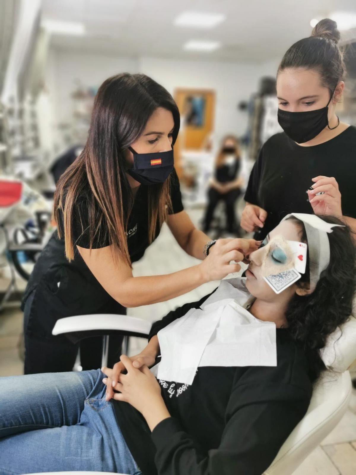 Tres mujeres, enmascaradas, en un salón. Una de ellas, tumbada con tarjetas sobre los ojos, recibiendo un procedimiento.