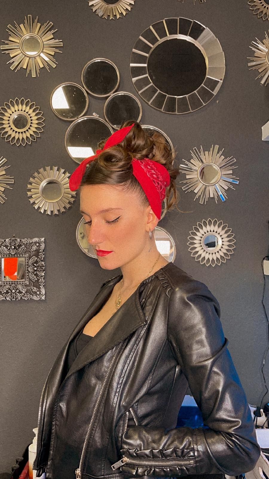 Mujer con chaqueta de cuero negra y pañuelo rojo en la cabeza, mirando hacia abajo, contra una pared con espejos.