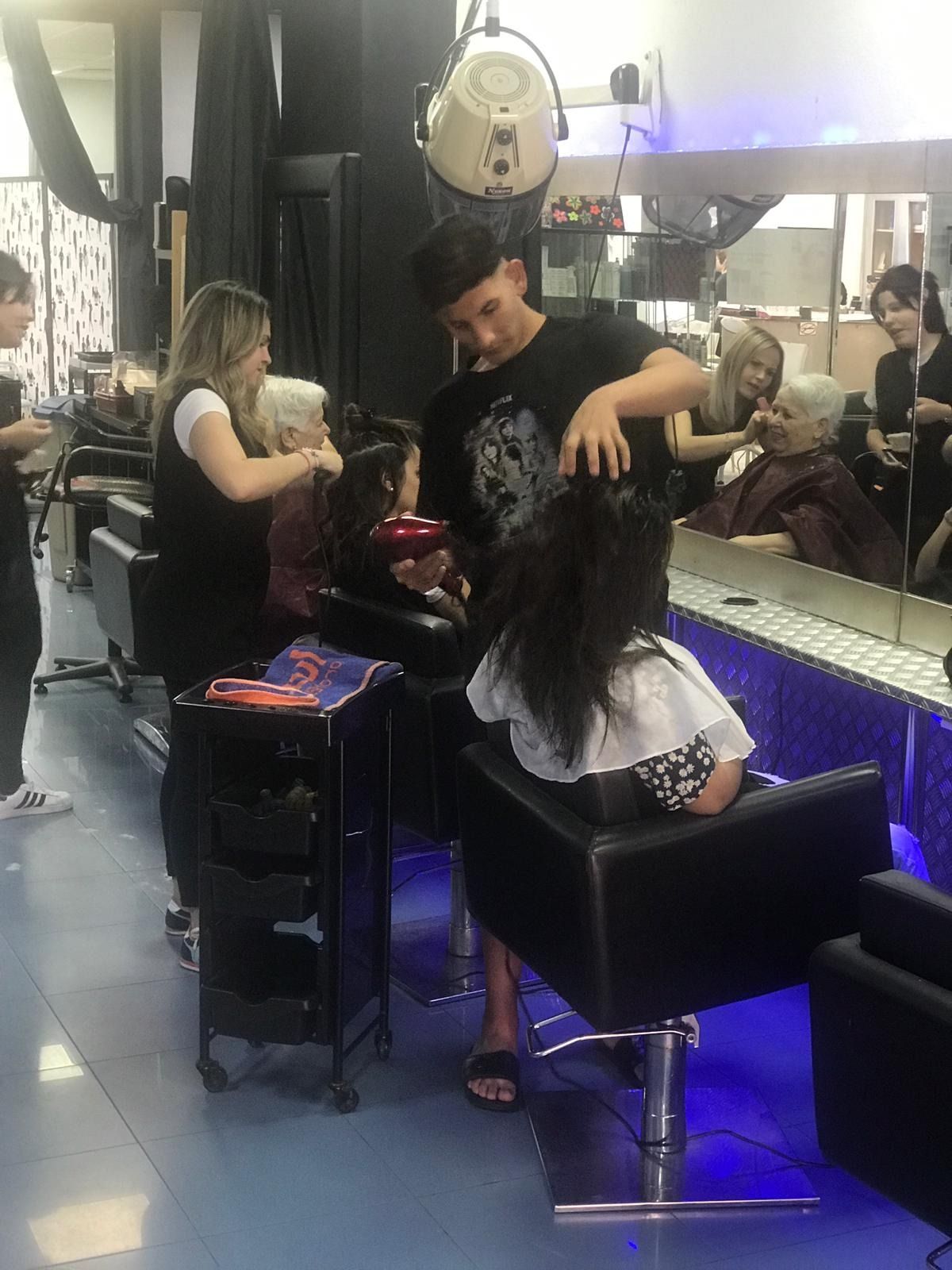 Estilistas trabajando con clientes en un salón; un hombre seca el cabello de una mujer sentada en una silla.
