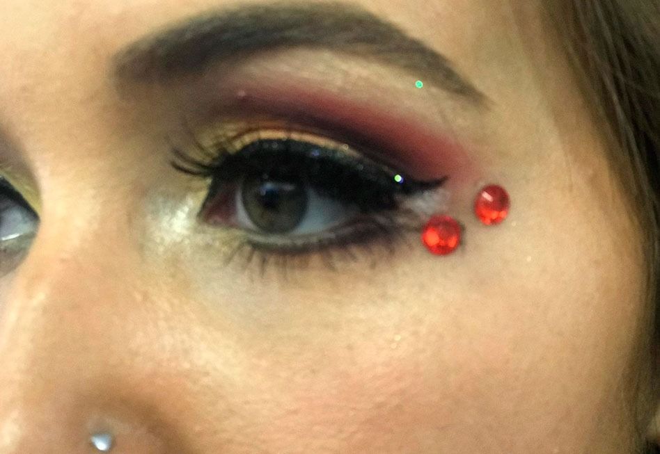 Primer plano de un ojo con maquillaje dramático: sombra de ojos roja, delineador de ojos alado, pestañas largas y gemas faciales.