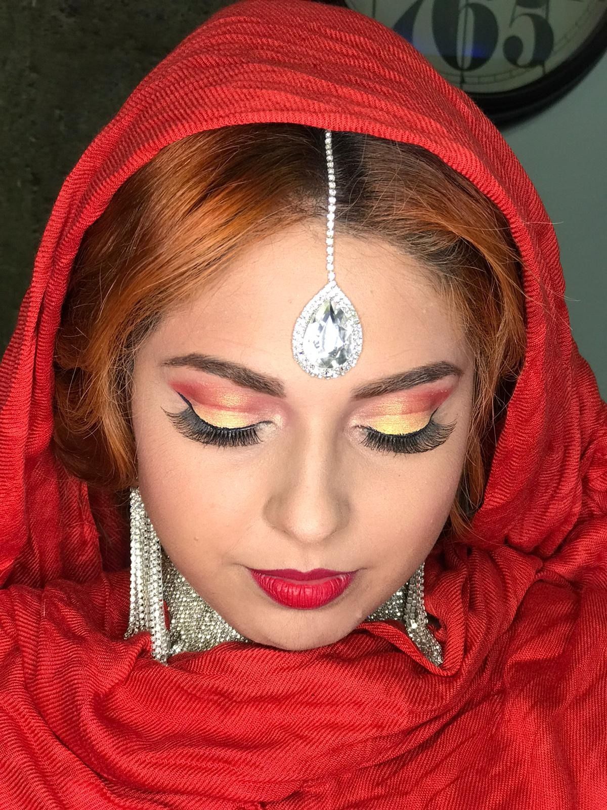 Mujer con hijab rojo y maquillaje dramático, incluido un bindi, mirando hacia abajo.