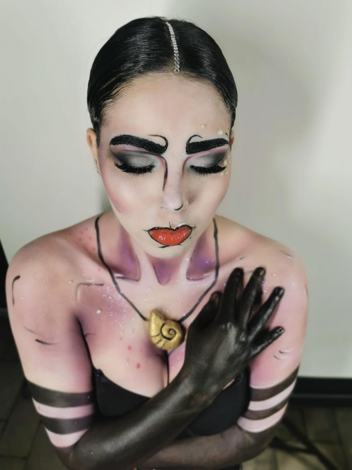 Mujer con maquillaje estilizado como Úrsula, brazos y torso negros, piel pálida, ojos cerrados, sosteniendo un collar de conchas.