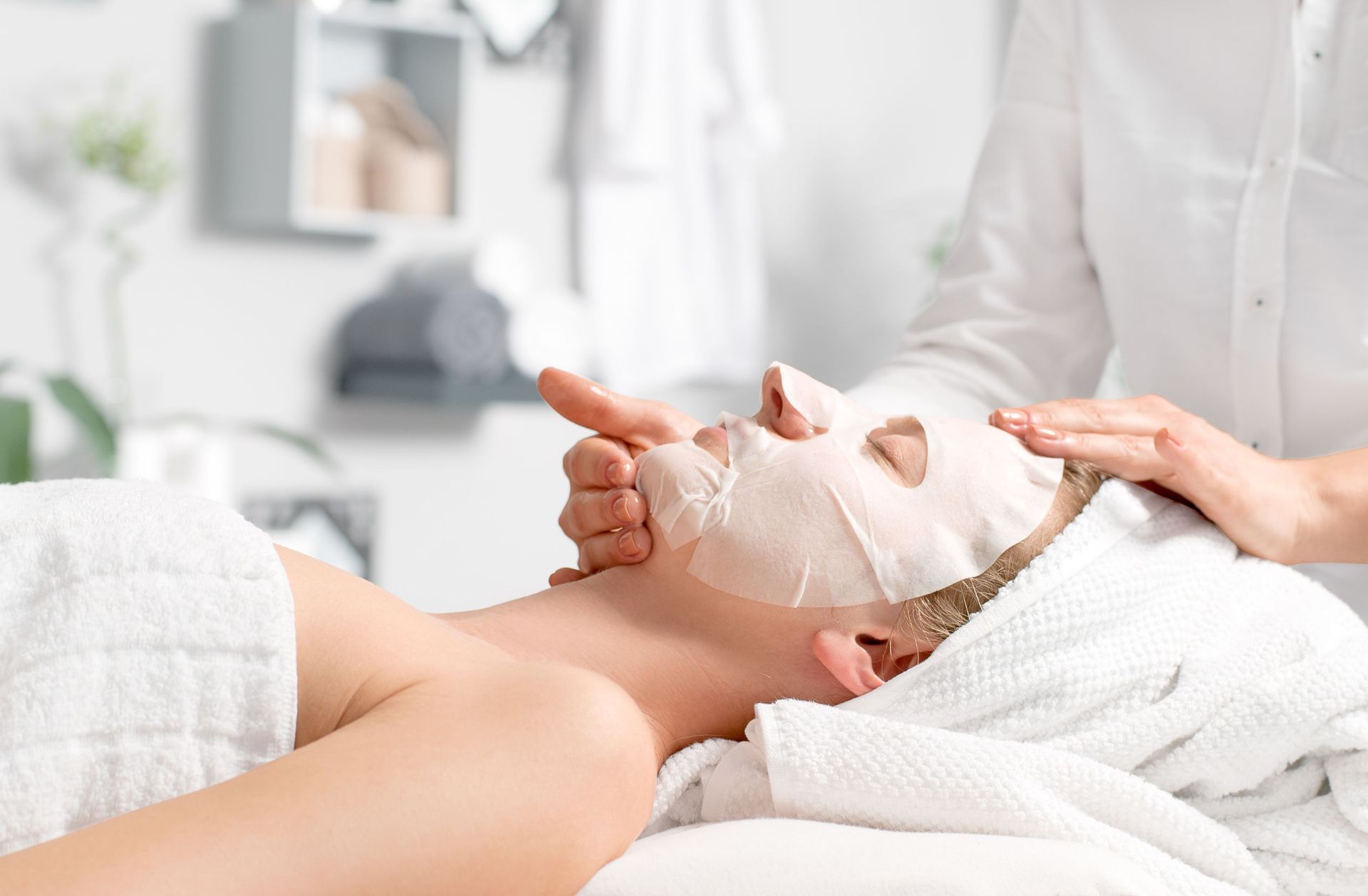 Mujer recibiendo mascarilla facial en un spa; manos del técnico alisando la mascarilla; bata blanca, fondo claro.