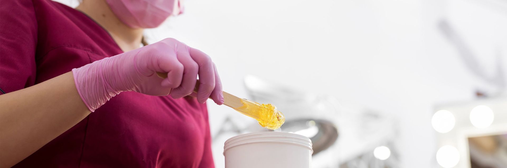 Persona con guantes y máscara de color rosa, sosteniendo una espátula amarilla con cera, sobre un recipiente blanco en un entorno luminoso.
