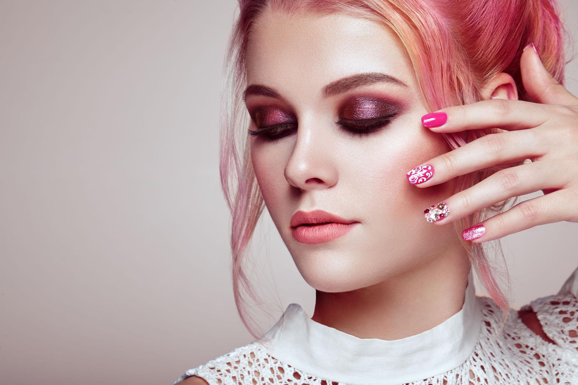 Mujer con cabello rosa, maquillaje de ojos y uñas estampadas tocándose la cara.