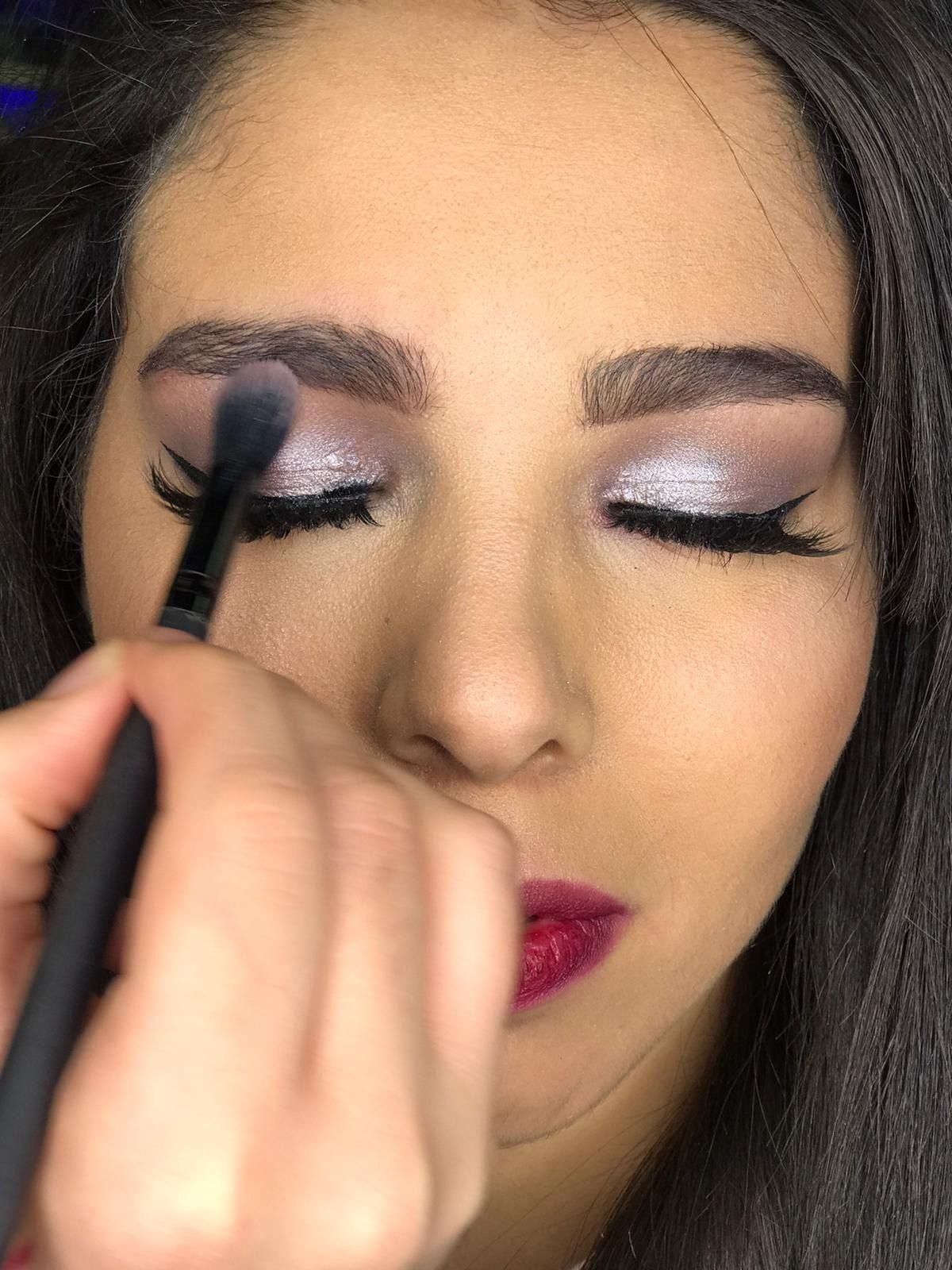 A una mujer se le aplica maquillaje de ojos con un pincel; tiene cabello oscuro, labios color ciruela y ojos cerrados.