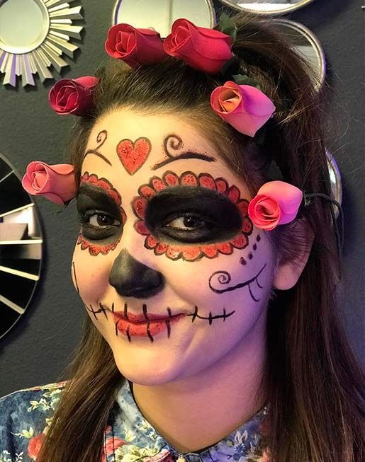 Mujer con maquillaje del Día de los Muertos y un accesorio para el cabello con una rosa sonríe. Ojos oscuros y camisa floreada.