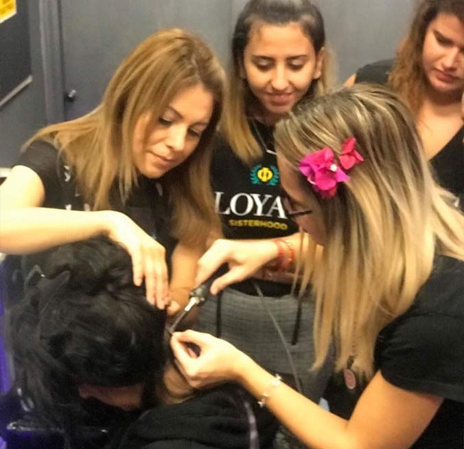 Cuatro mujeres peinando a una persona, probablemente extensiones. Interior. Expresiones concentradas.