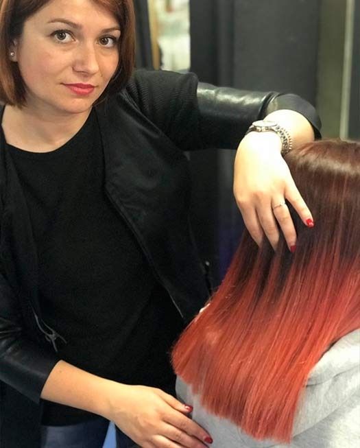 Una mujer peinando el cabello ombré rojizo de una clienta en un salón. Lleva una chaqueta negra y mira a la cámara.