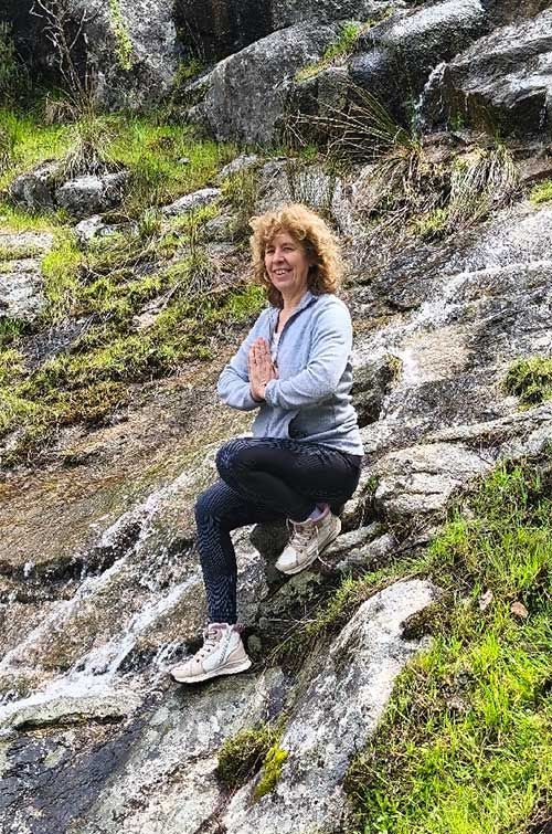 Mujer en pose de yoga en una ladera rocosa, con los brazos en oración, sonriendo.