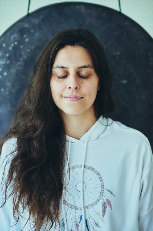 Mujer de pelo largo y oscuro, con ojos cerrados, sonriendo, delante de un gran gong, vestida con una sudadera con capucha blanca.