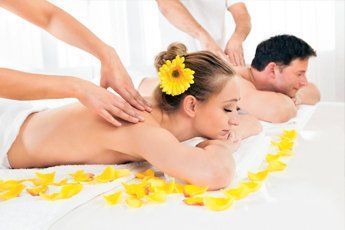 Pareja recibiendo masajes en spa, acostados boca abajo, flor amarilla, pétalos en la cama.