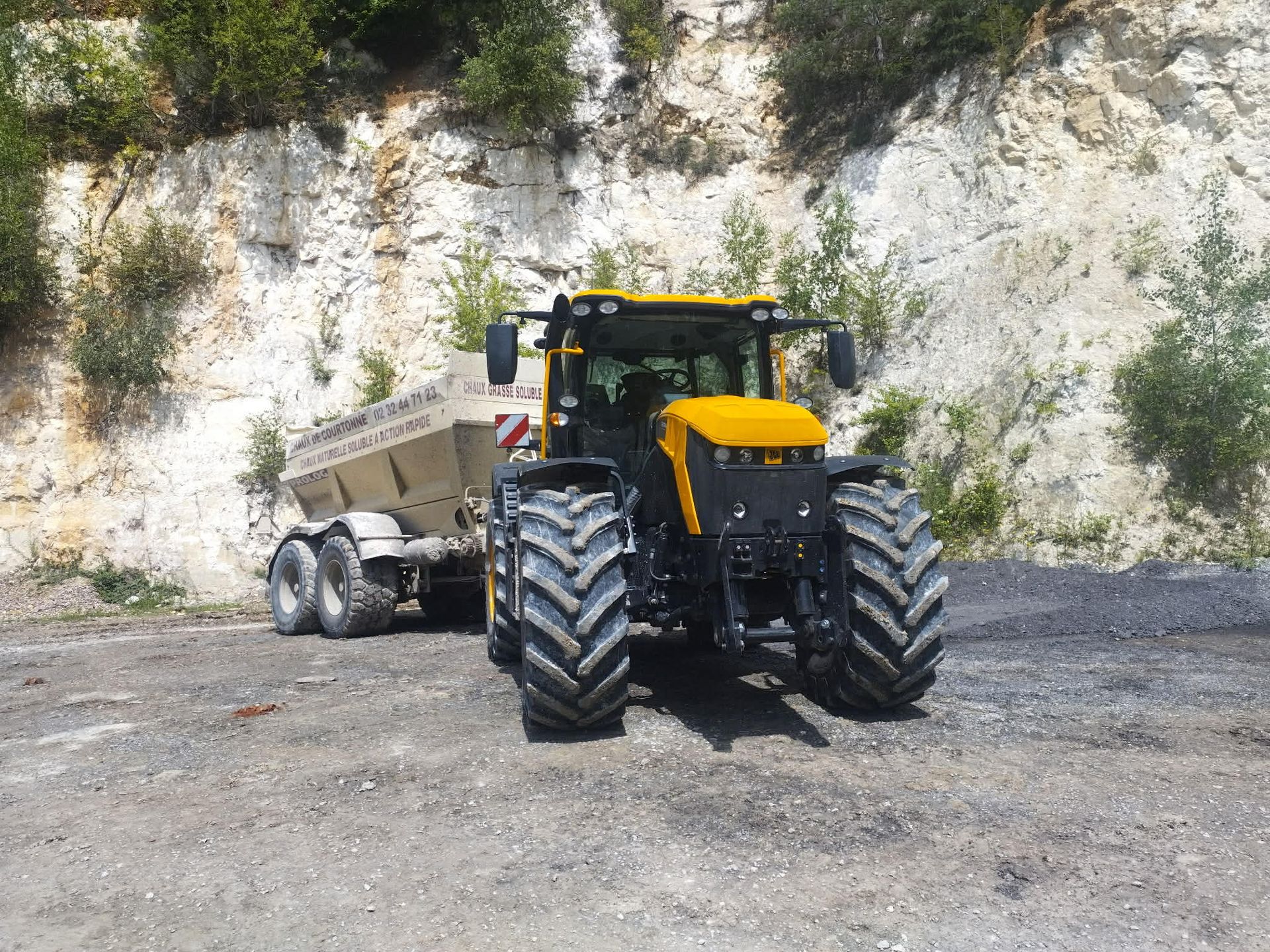 Un tracteur jaune et noir tirant une remorque chargée de terre devant une falaise rocheuse claire.